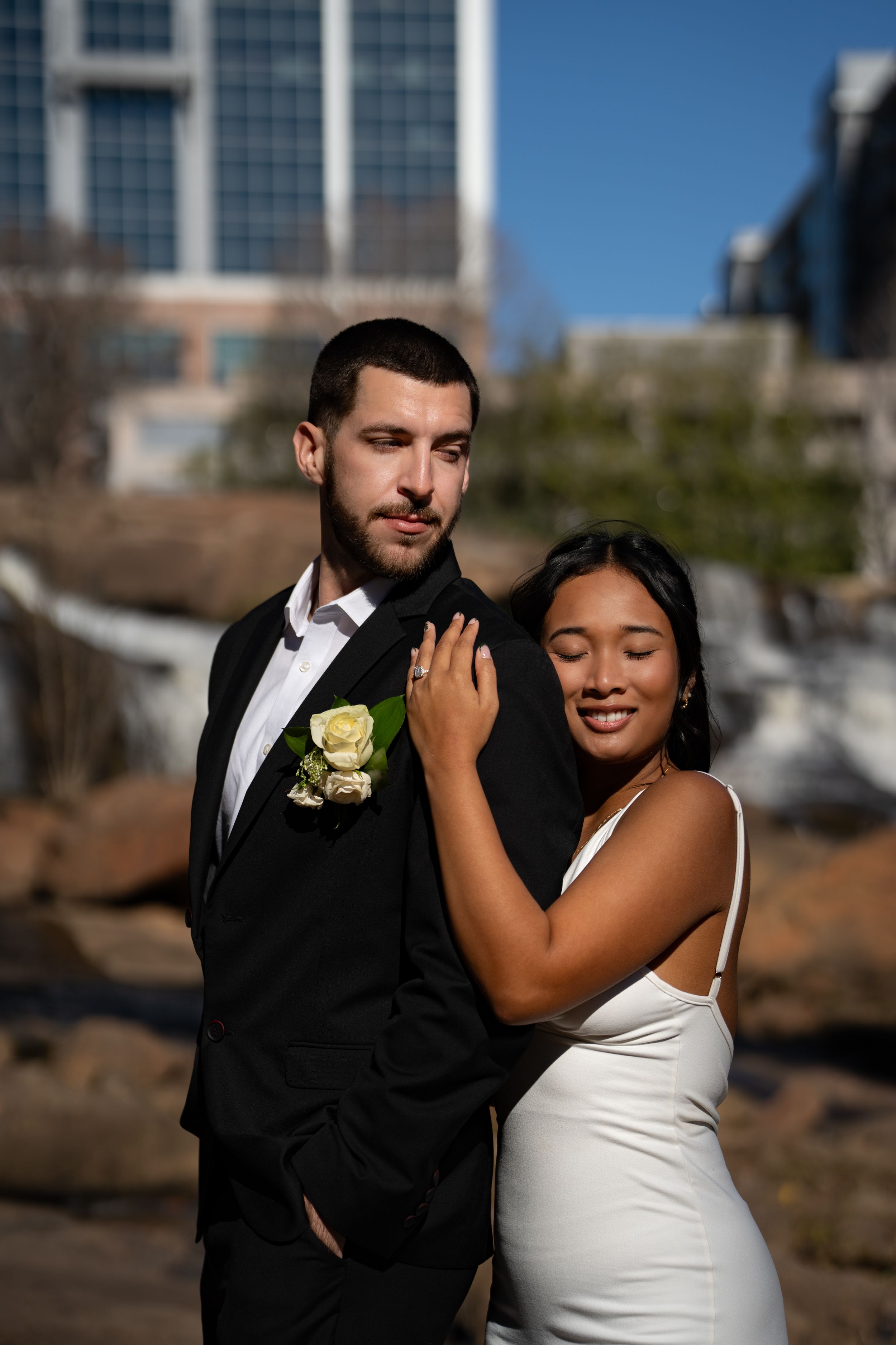 A couple in formal wedding attire share intimate moments during an outdoor photo session against a city backdrop.