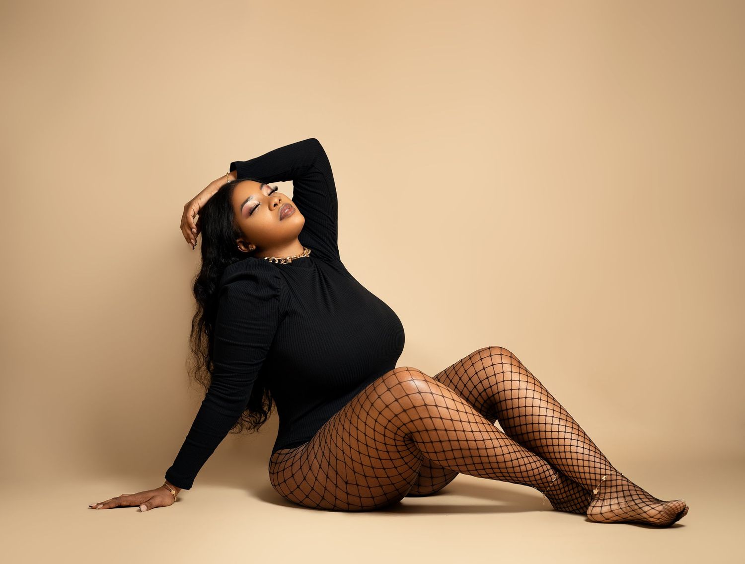 Fashion model in black top and fishnet stockings poses on beige background in studio setting.