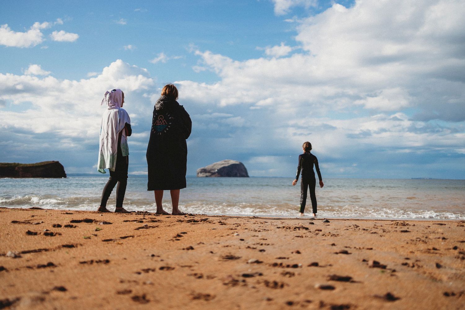 Three silhouettes standing on a sandy beach looking out at the ocean and rocky islands.