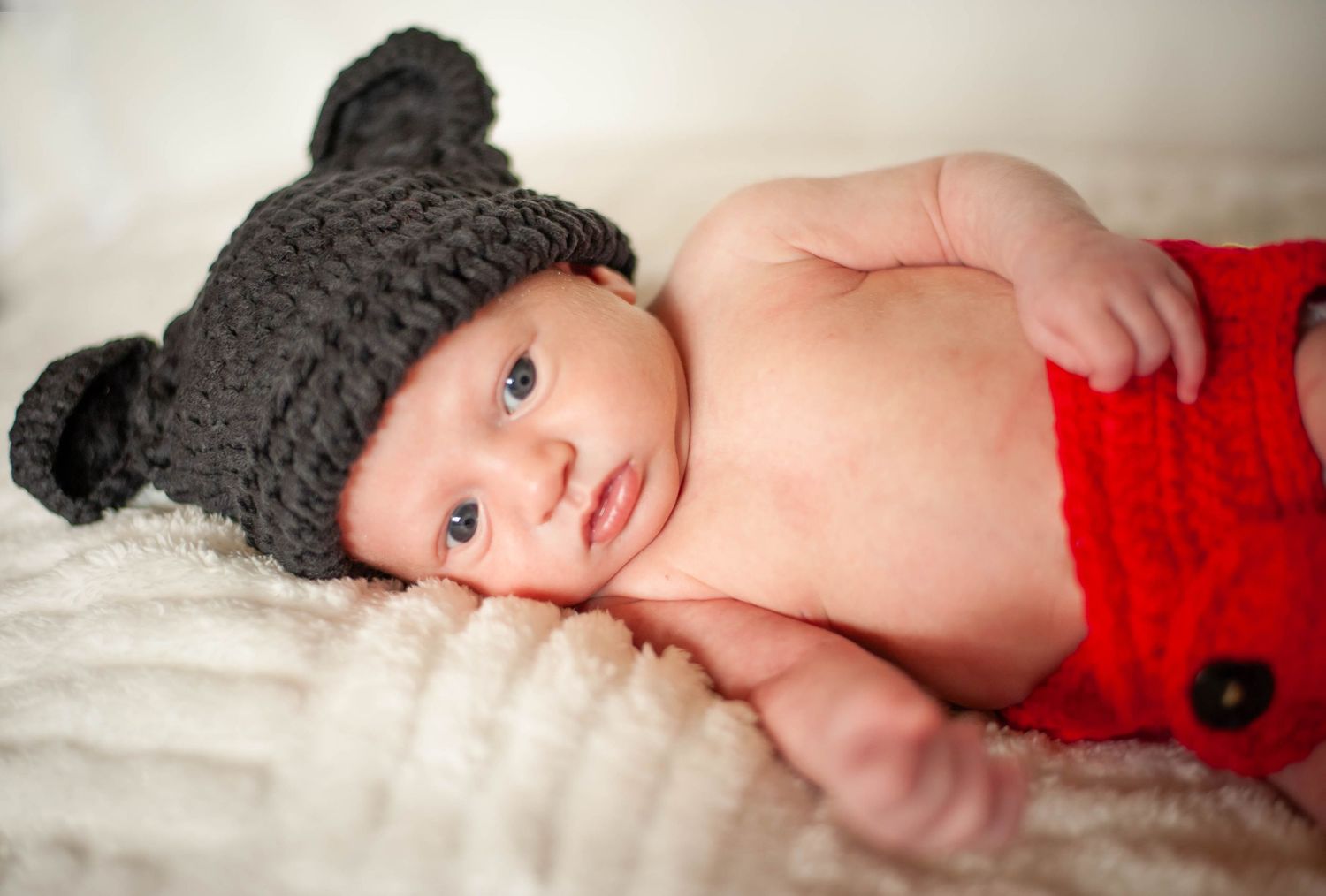 Baby wearing knit bear hat and red bottoms laying on white blanket.