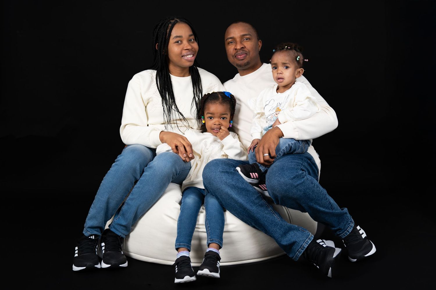 A family of four wearing white tops and blue jeans poses together on a white chair against a black backdrop.