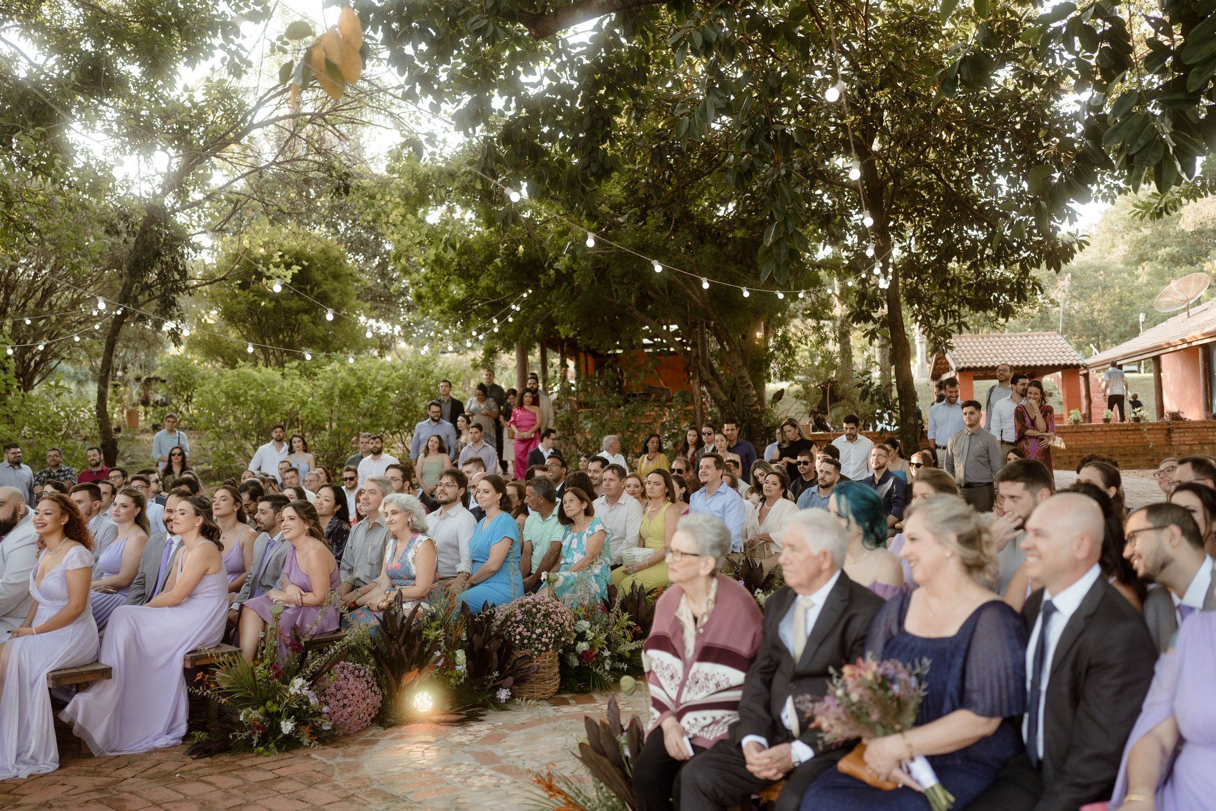 Wide view of wedding guests seated outdoors under string lights and tree canopy.