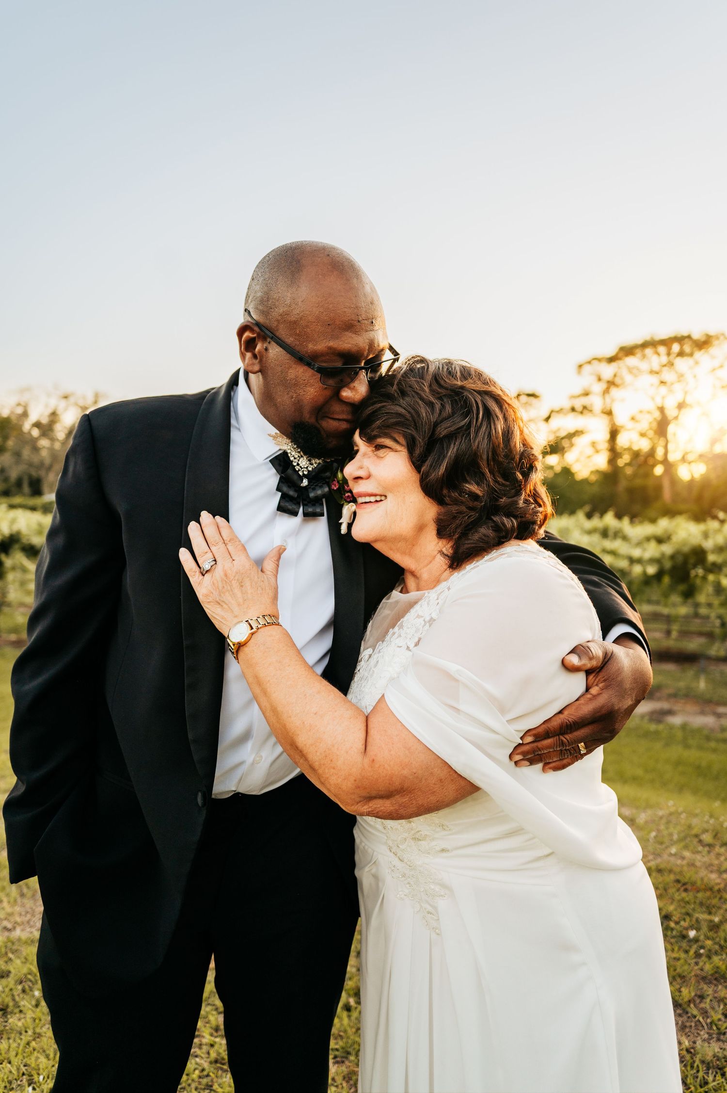 Intimate moment between newlyweds in formal attire against a sunset vineyard backdrop.