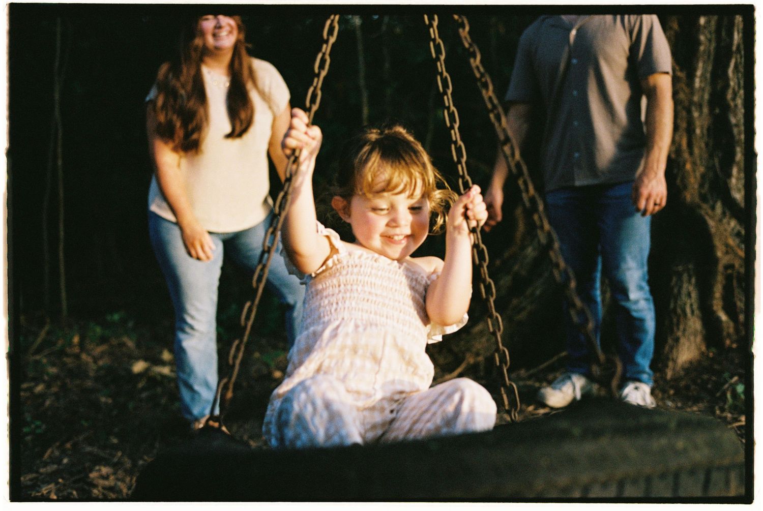 A family pushing their toddler on a tire swing in a forest setting, shot on vintage film.