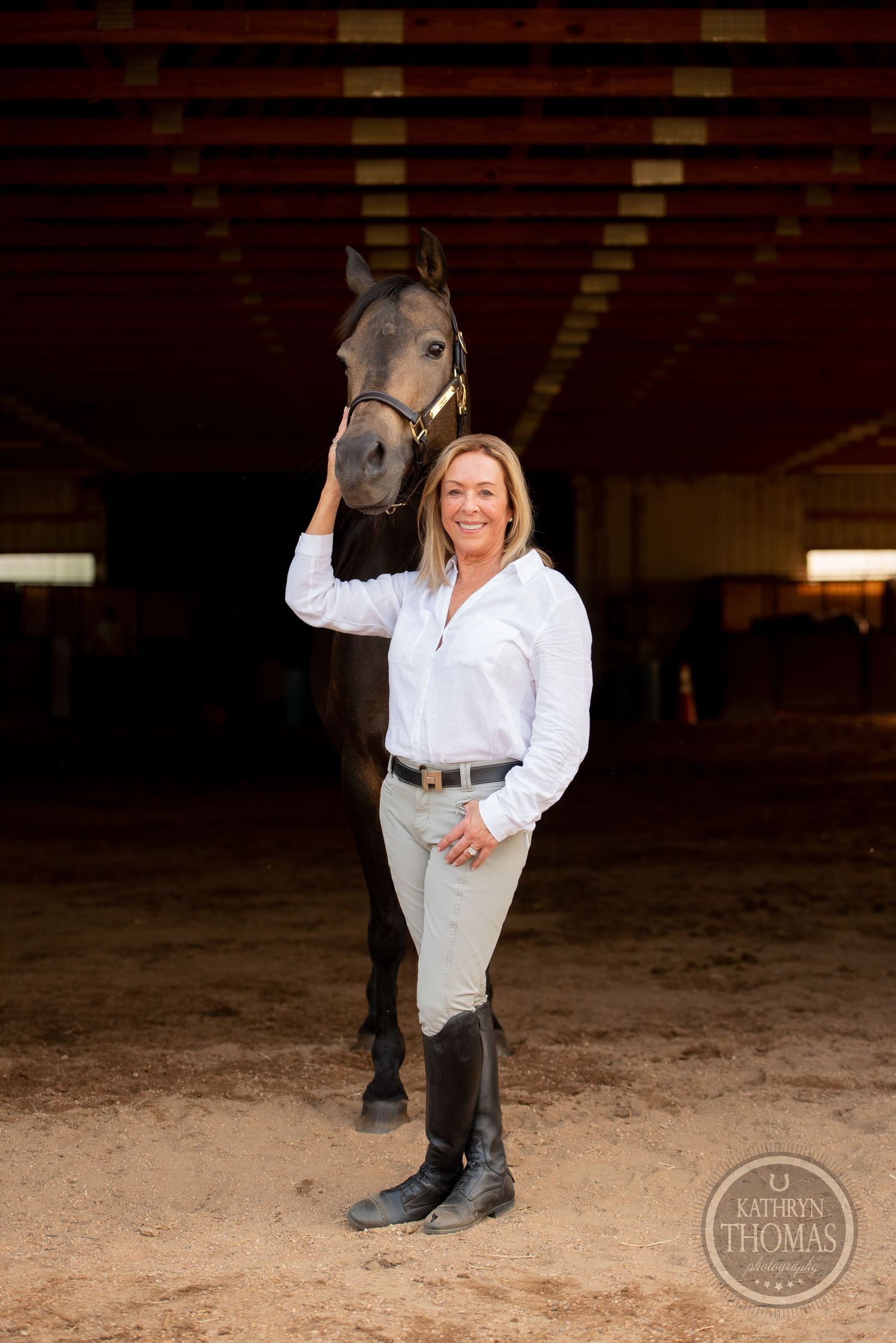 Equestrian in white shirt and riding boots poses with horse in indoor arena setting.