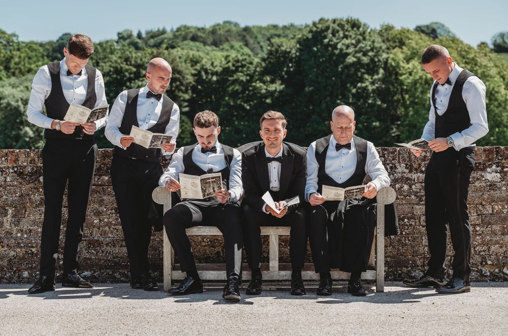 Groomsmen in matching black vests and bowties pose on stone wall at outdoor wedding venue.