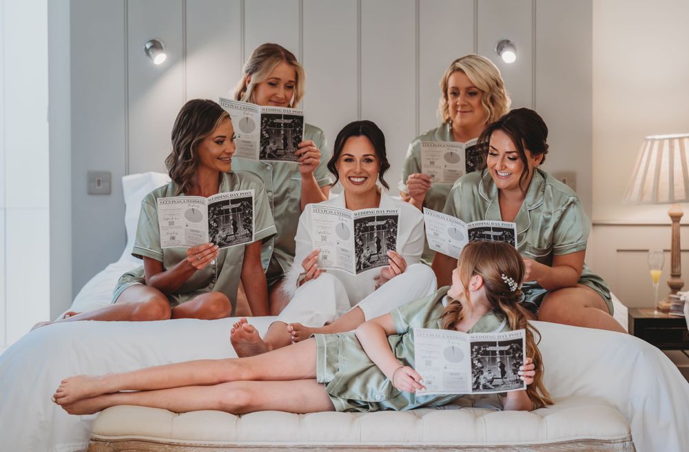 Bridesmaids in pastel robes relaxing on bed while reading papers together.