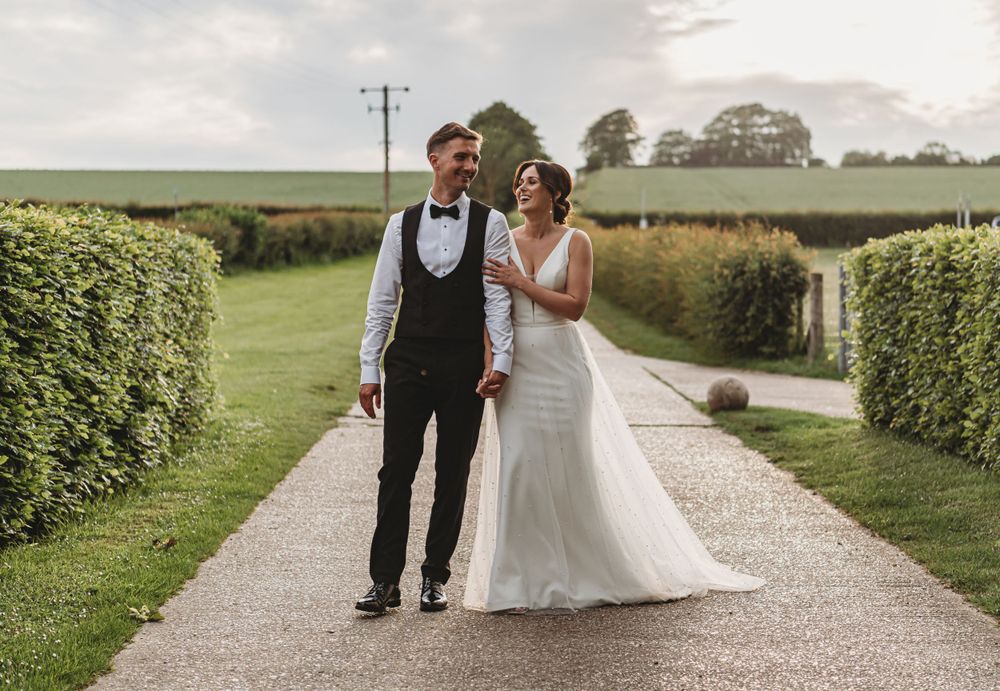 A wedding couple walks together on a manicured garden path surrounded by hedges.