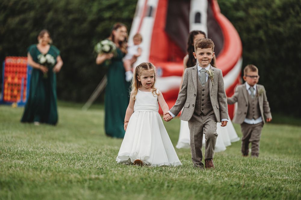 Young attendants in formal wear walk on grass with an inflatable slide visible in background.