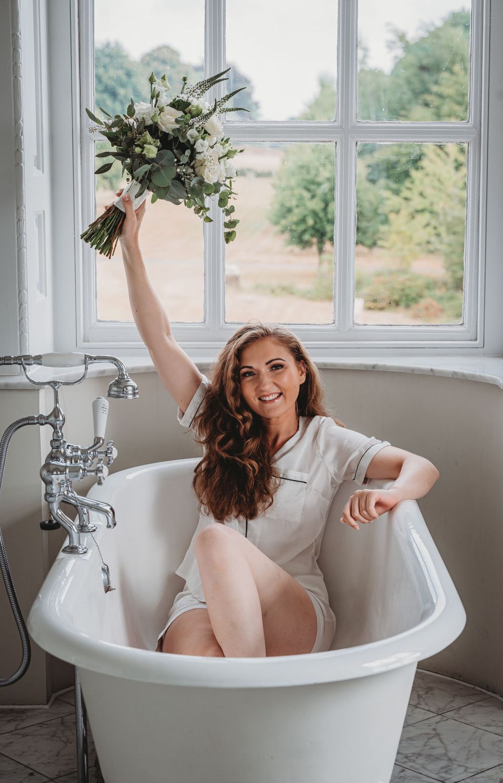 Playful bridal portrait in vintage clawfoot tub holding white bouquet.