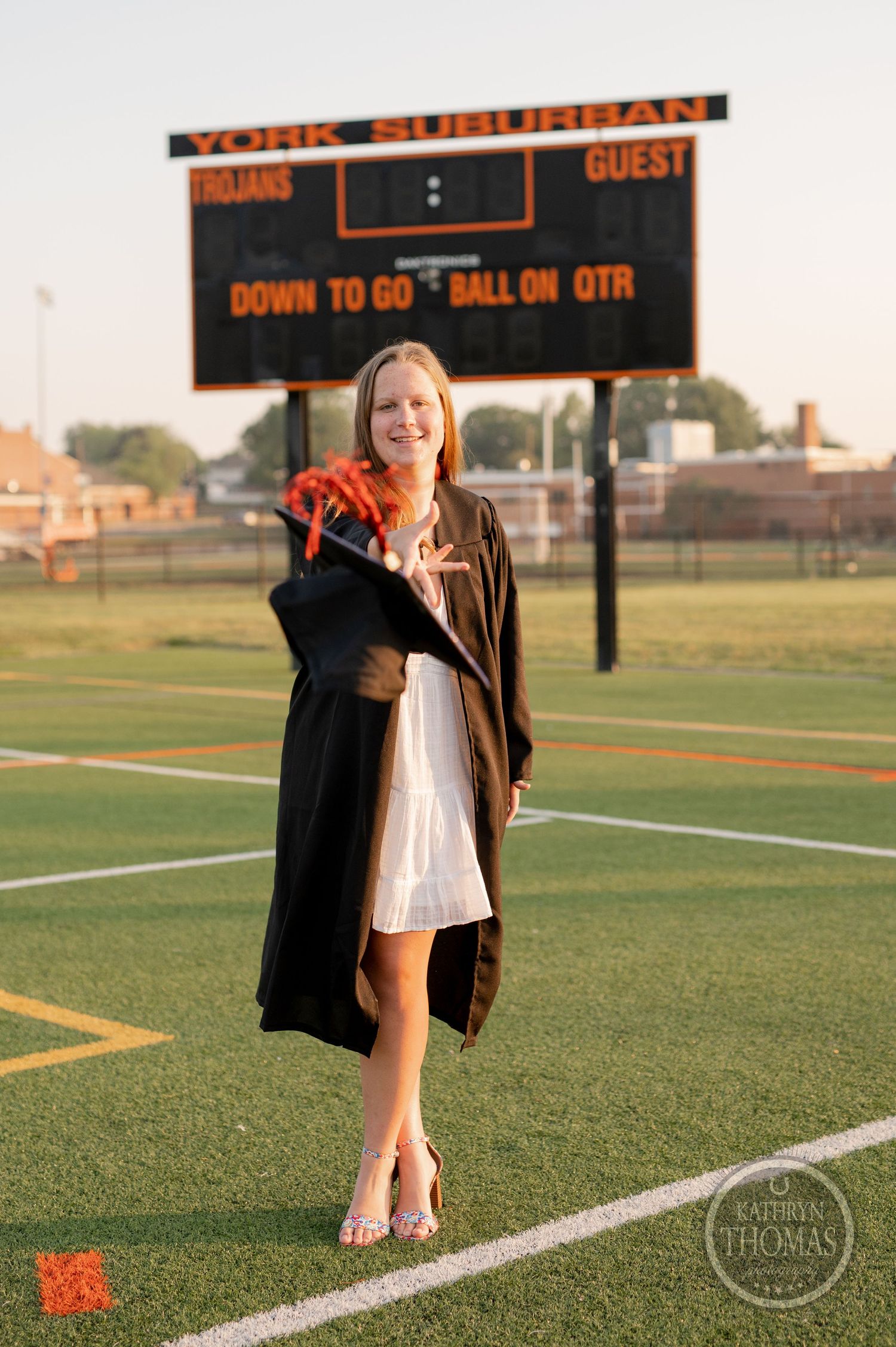 Graduate in white dress and black gown standing on football field with scoreboard in background.