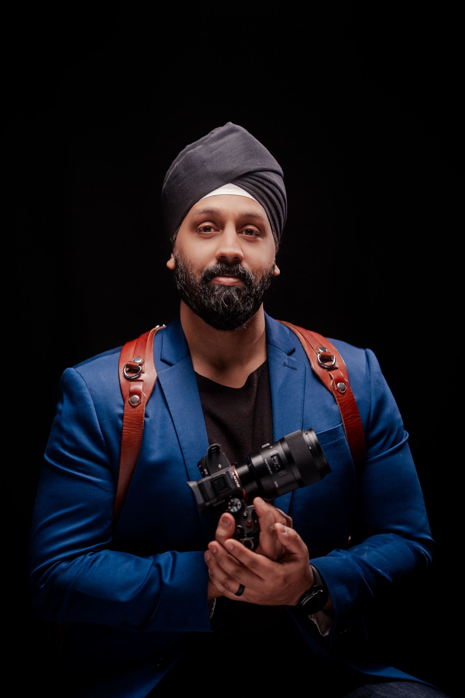 Professional photographer in a blue blazer and turban holding a camera against a black background.