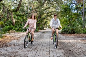 Two people riding bicycles down a scenic tree-lined brick path surrounded by lush tropical greenery.