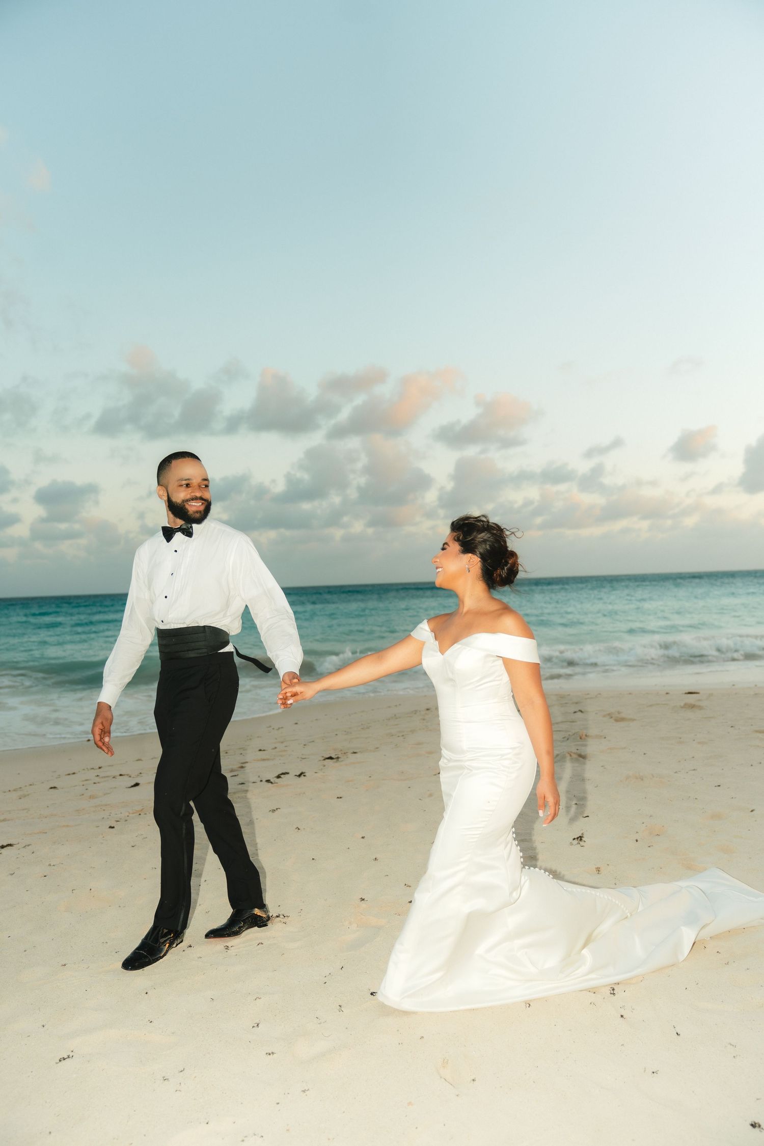 Playful couple dances on a pristine beach at sunset with flowing white dress.