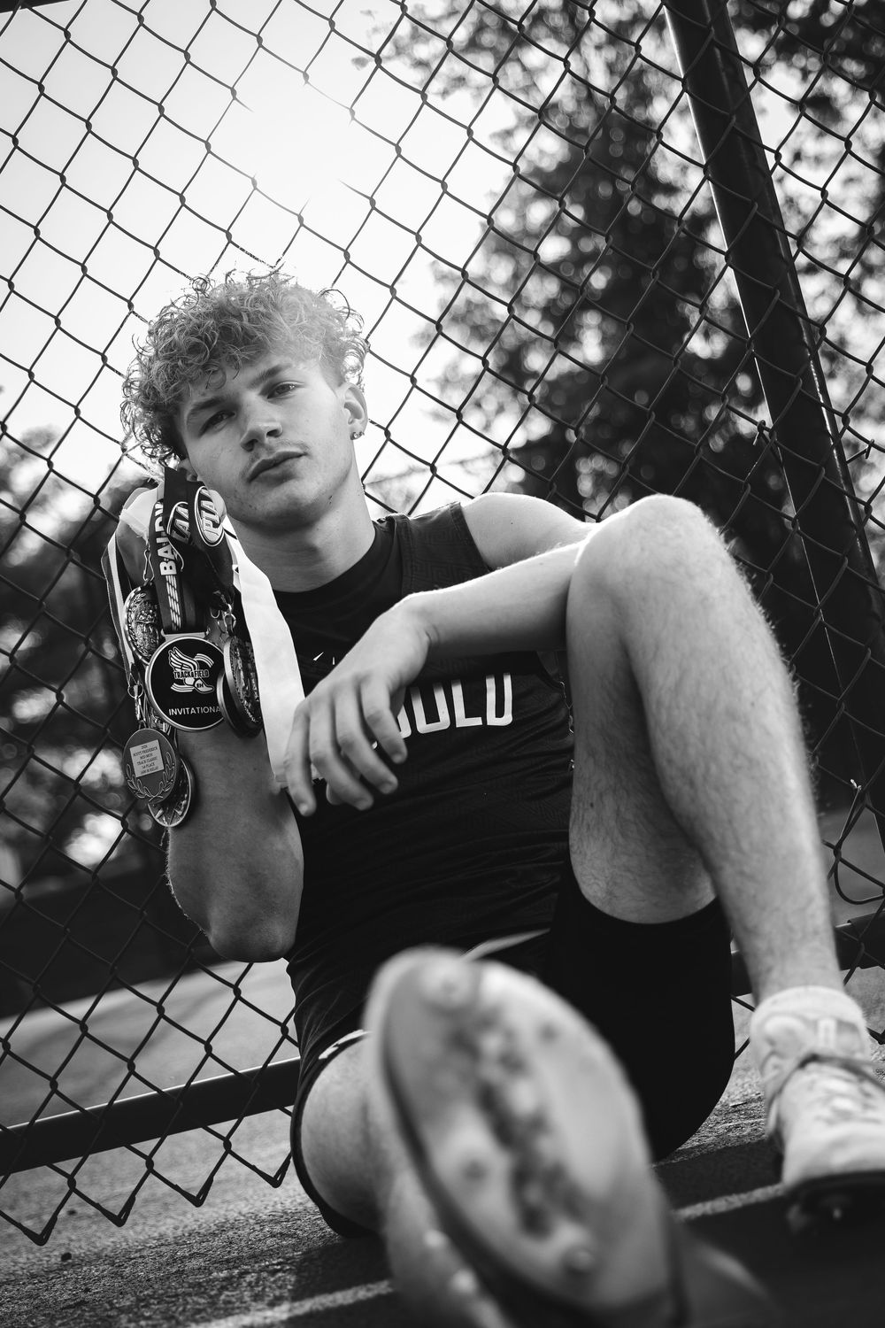 Young male athlete sitting against chain-link fence holding track medals in black and white photo.