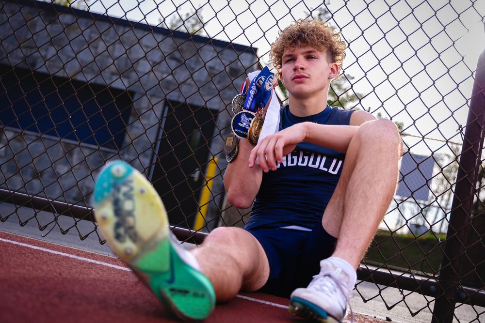 Young male track athlete sitting against chain-link fence holding trophy wearing navy uniform and green spikes.