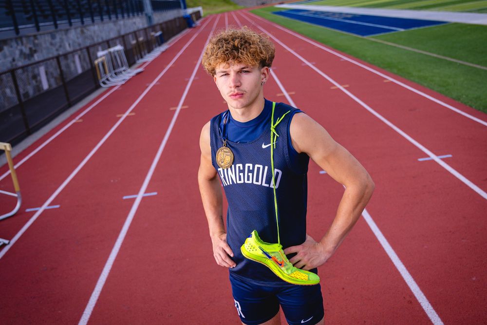 Young male track athlete in Ringgold uniform holding yellow spikes on a red running track.