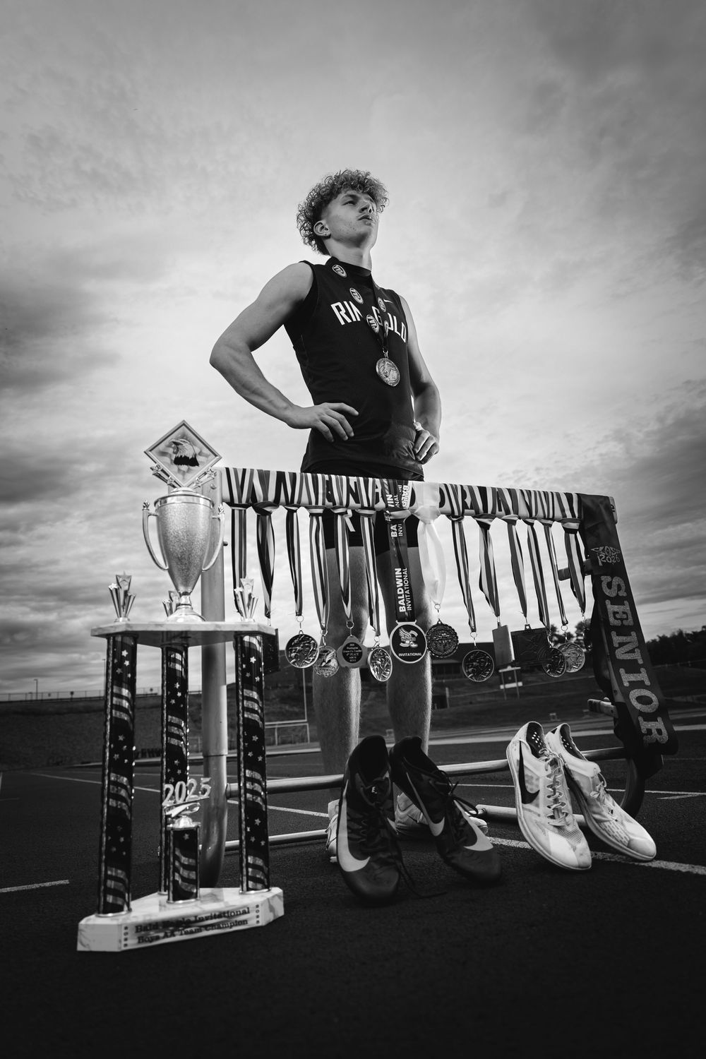 Black and white photo of a young male athlete standing proudly with trophies and medals on a track.