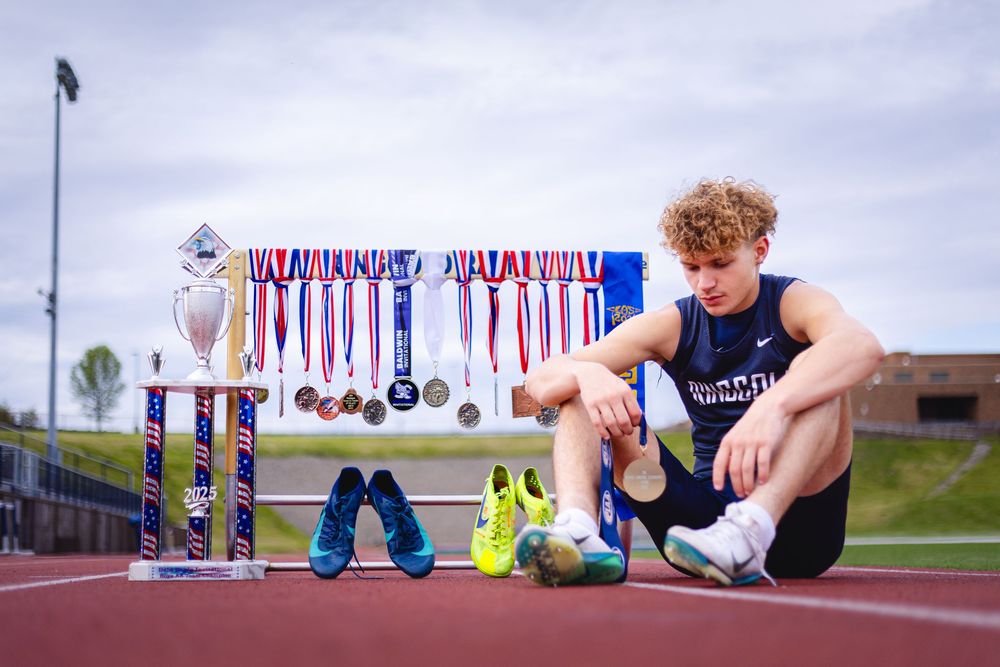 Young male track athlete sits on a track surrounded by trophies, medals, and colorful racing spikes.