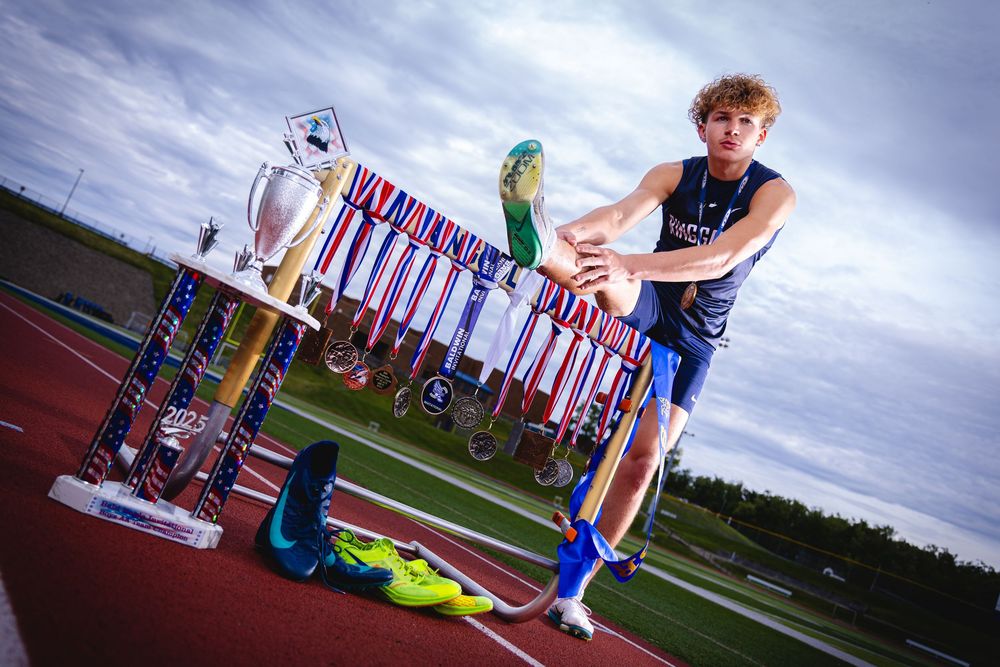 Young male track athlete stretching beside trophy and medals on a running track, wearing blue athletic wear.