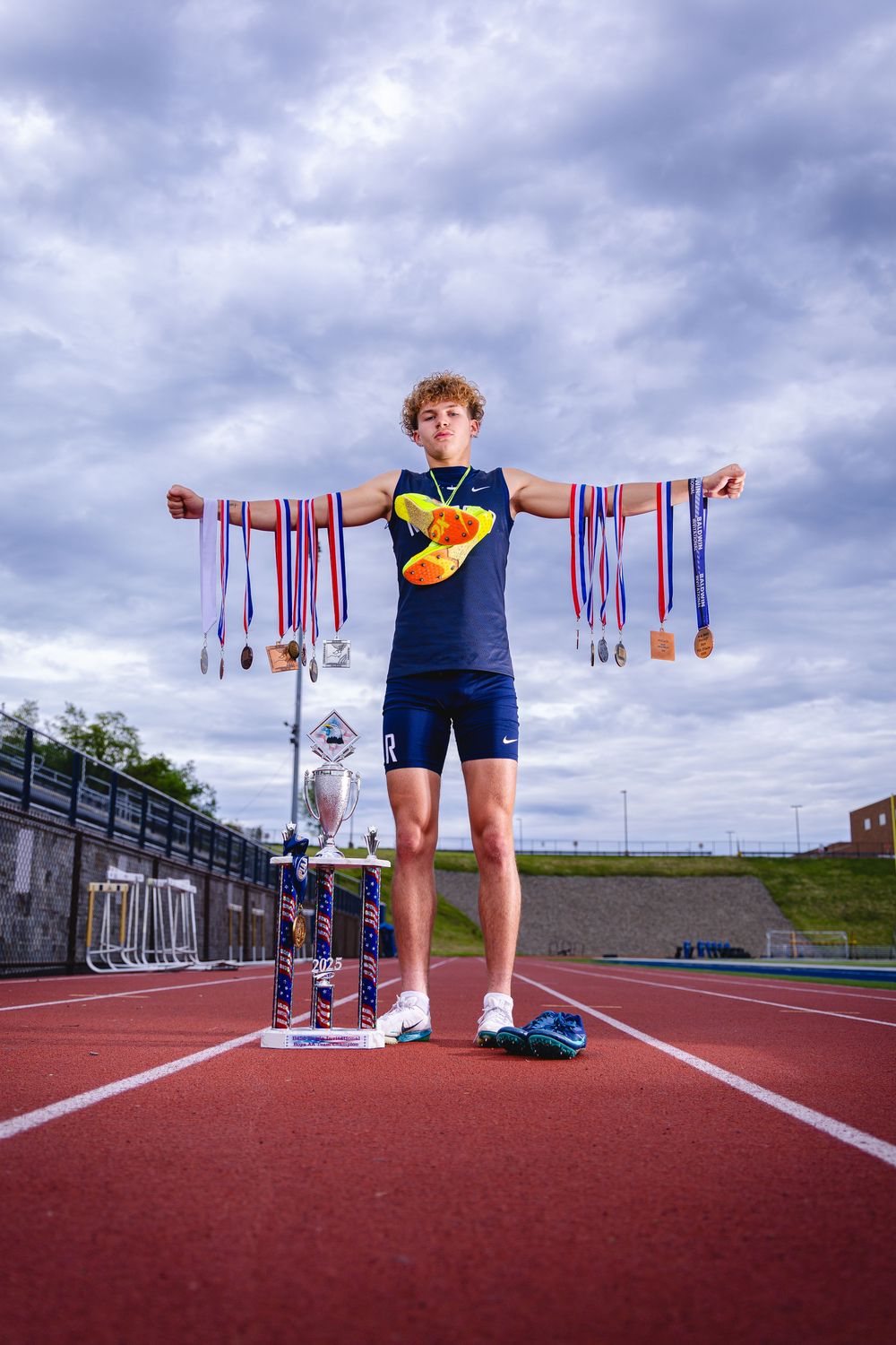 Young male track athlete stands on a red track displaying numerous medals with arms outstretched and a trophy at his feet.