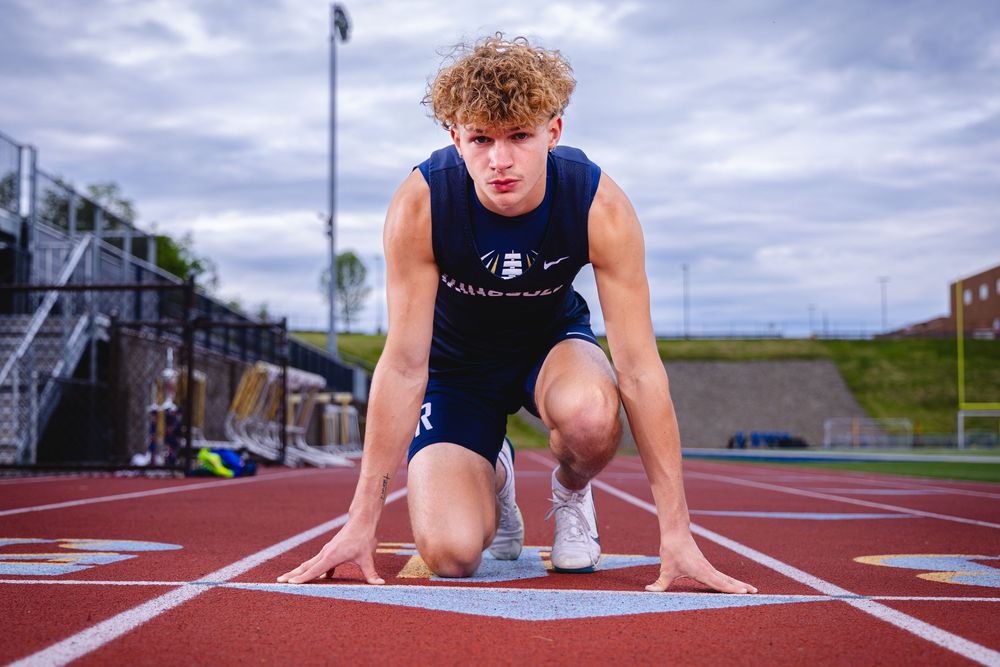 Young male sprinter in starting position on red track wearing navy blue athletic uniform under cloudy sky.