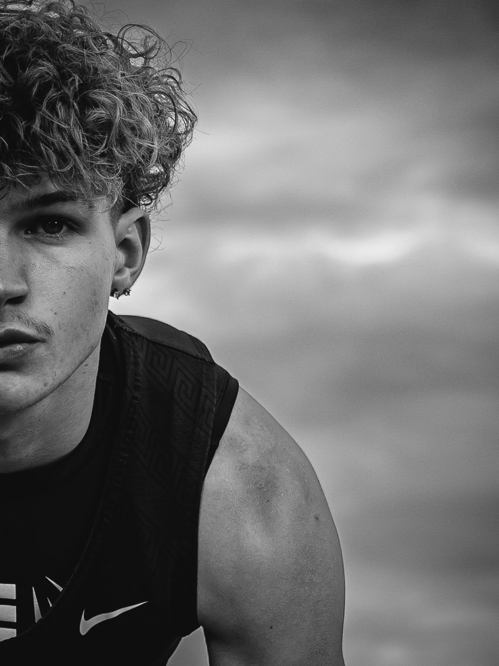 Black and white portrait of a young athletic male with curly hair wearing a Nike sleeveless shirt outdoors.