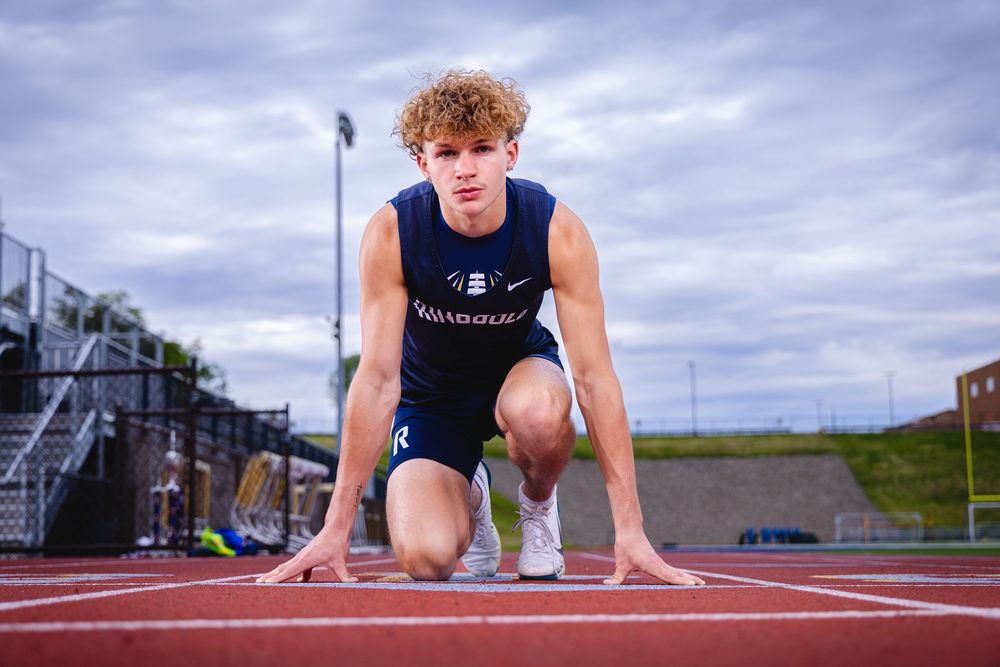 Teen track athlete crouched in starting blocks on red running track wearing navy Ridgould uniform.