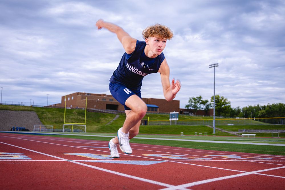 A young male sprinter explodes off the starting line on a red track wearing a navy blue athletic uniform.