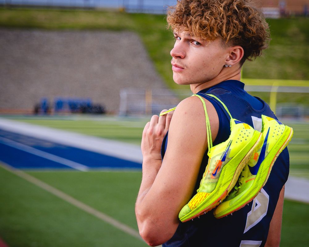 Young male athlete holding neon yellow Nike track spikes on a blue stadium field.