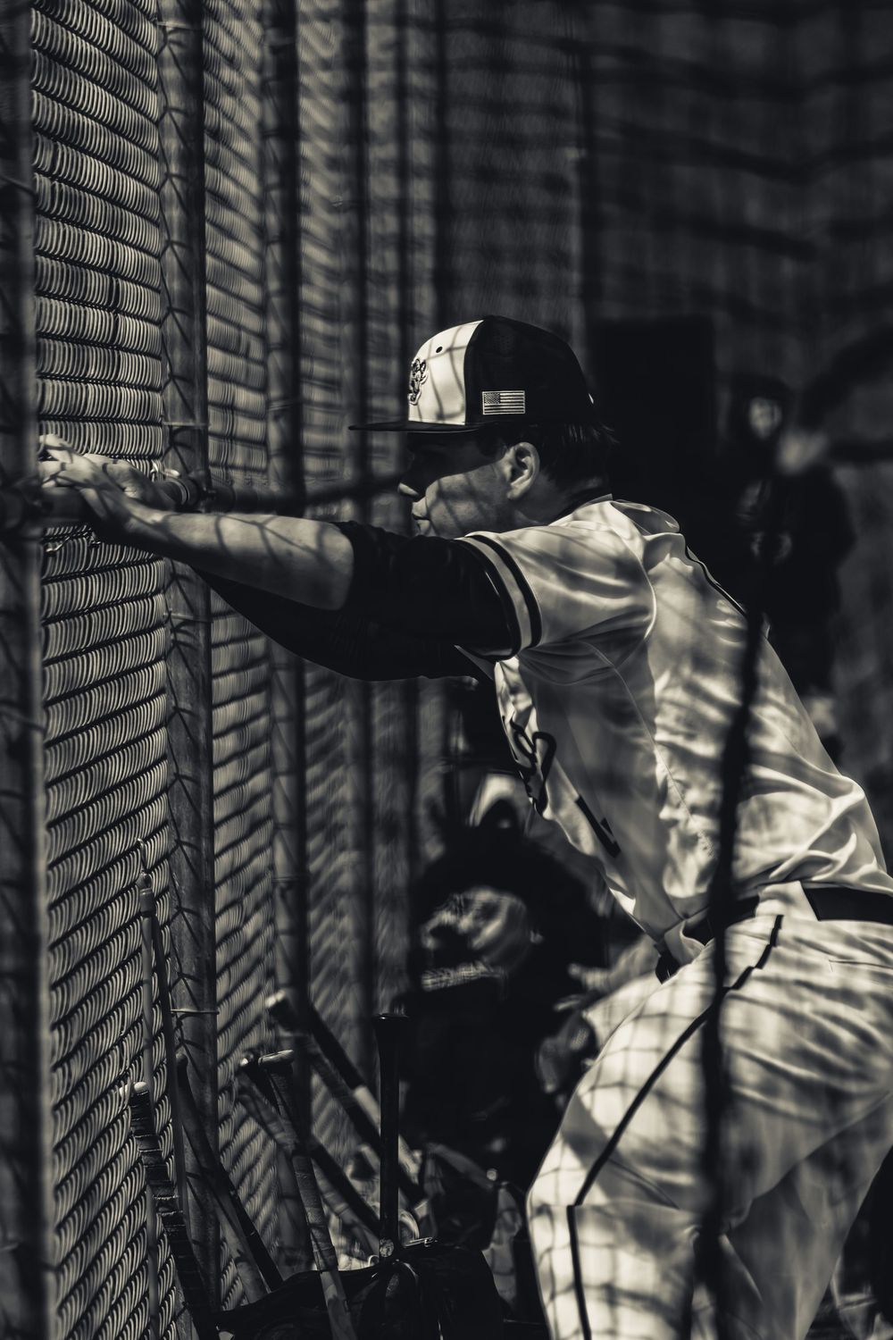 Black and white photo of a baseball player leaning against a batting cage net during practice.