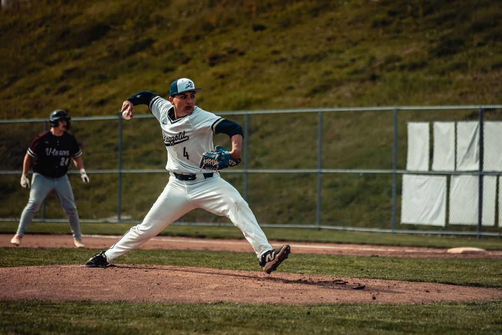 Baseball pitcher in green uniform mid-throw on field, with baserunner and opponent visible in background.