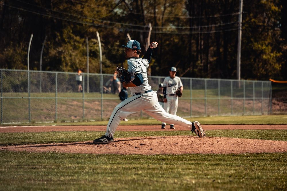 Baseball pitcher in mid-throw on a sunny day, wearing a teal and white uniform on a well-maintained diamond.
