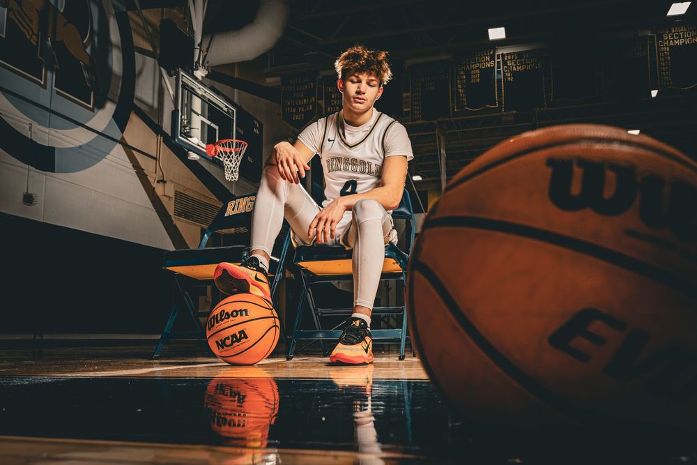 Basketball player sits on wooden court with Wilson basketballs in dramatic lighting at indoor gymnasium.