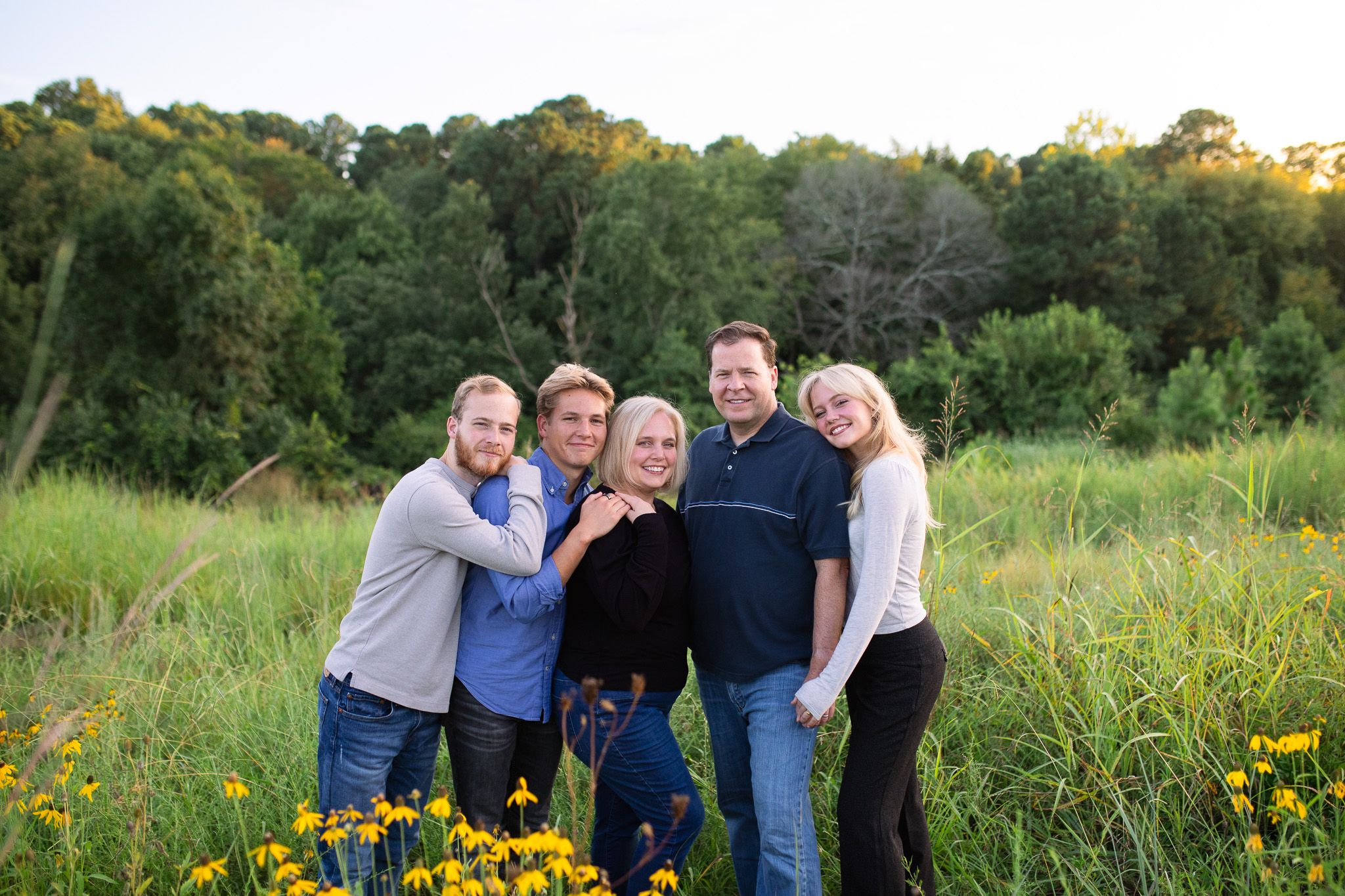 A group stands together in a meadow with yellow wildflowers and green trees in the background during golden hour.