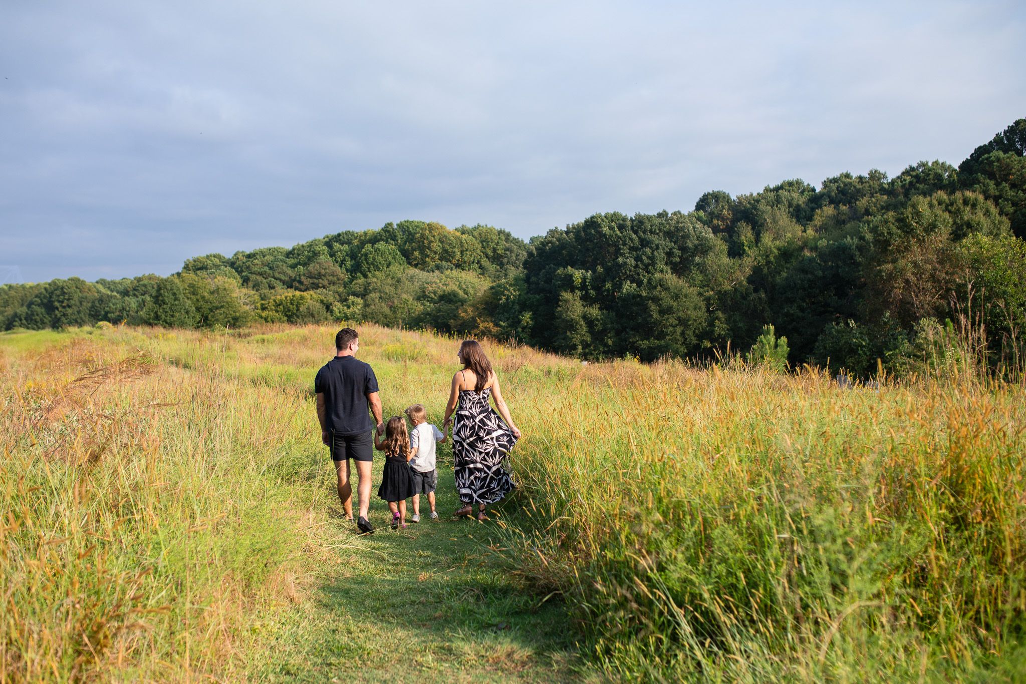 Two people and a dog walking together on a dirt path through a golden meadow with green forest in the background.