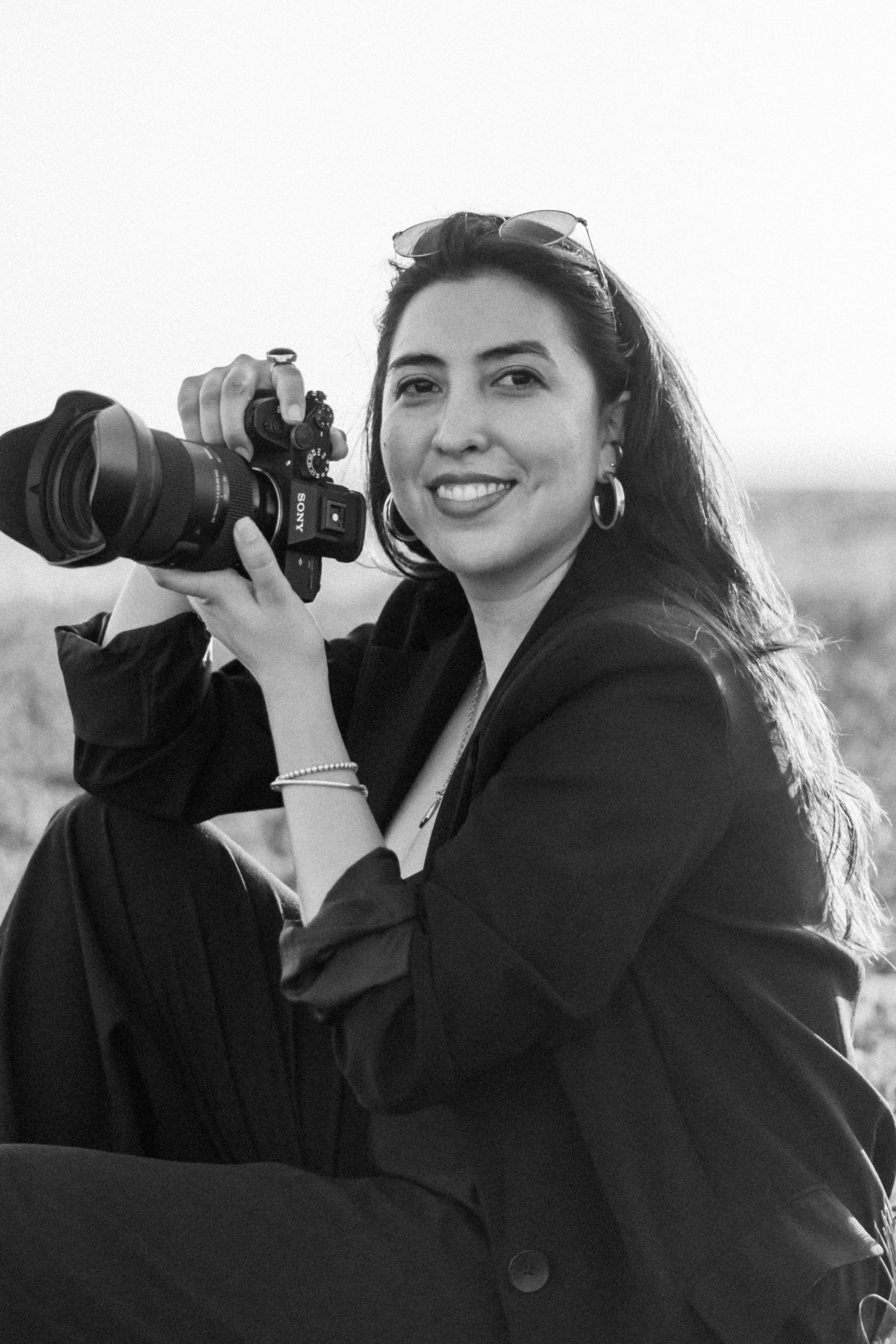 Photographer with long dark hair holds camera while smiling outdoors.