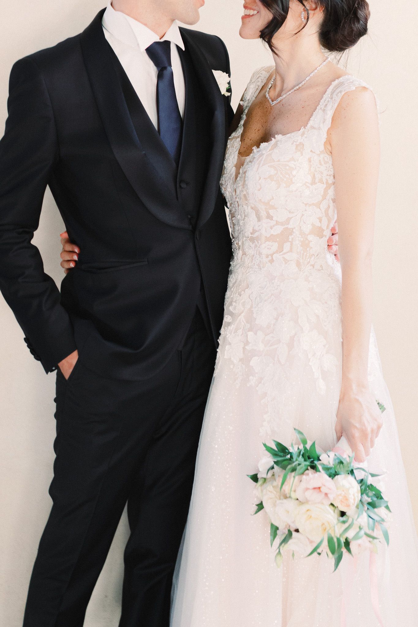 elegantly dressed bride and groom, smiling, hugging, holding a wedding bouquet
