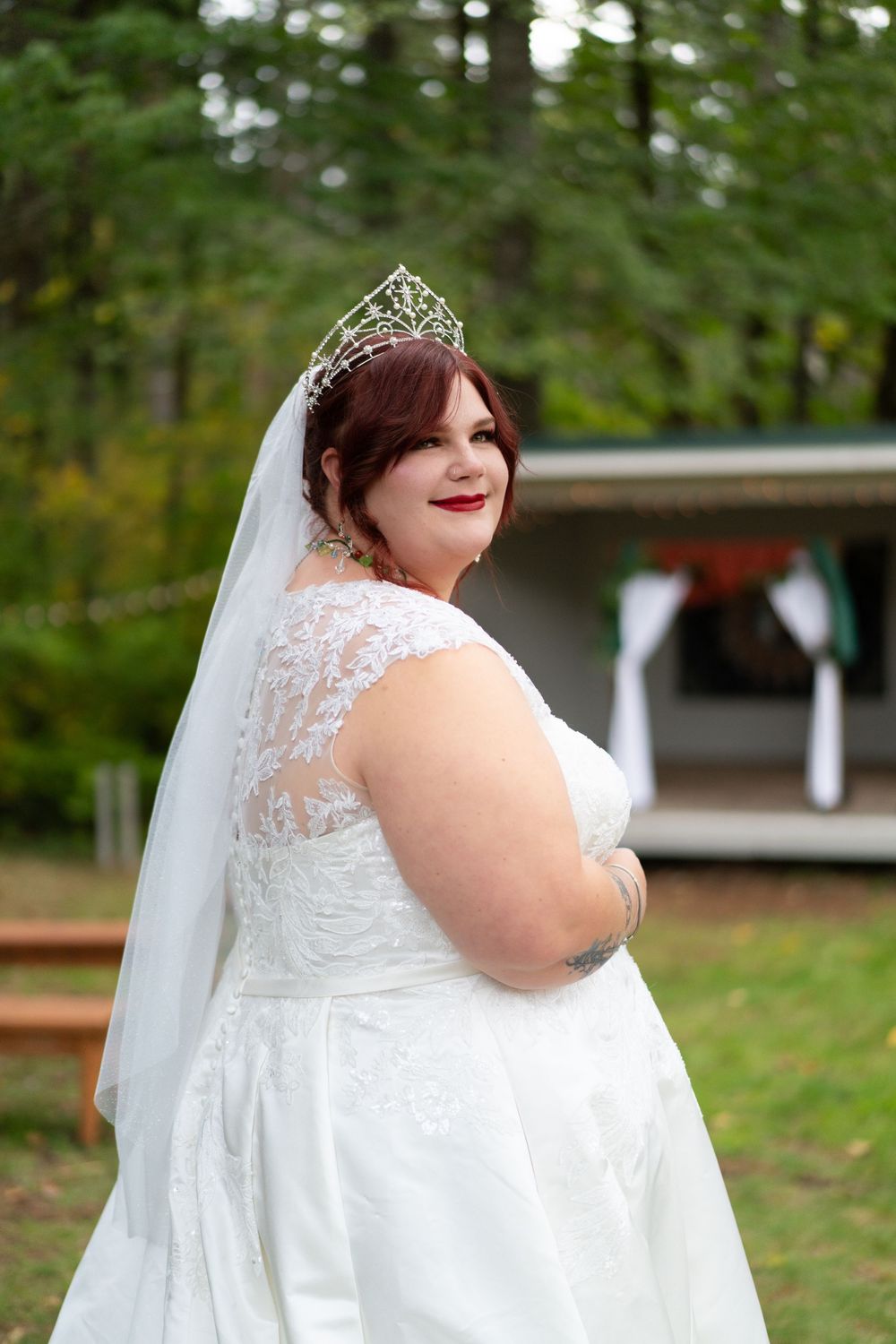 A joyful bride in a white lace wedding dress poses outdoors in a natural setting with trees in the background. captured by Savvy Photography