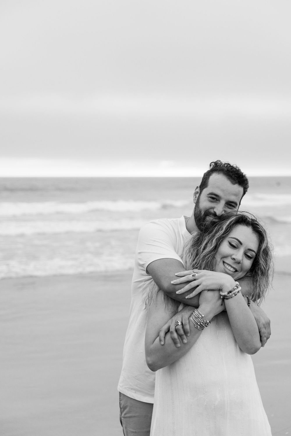Candid moment of affection between a couple, captured in black and white on a misty beach by an engagement photographer.