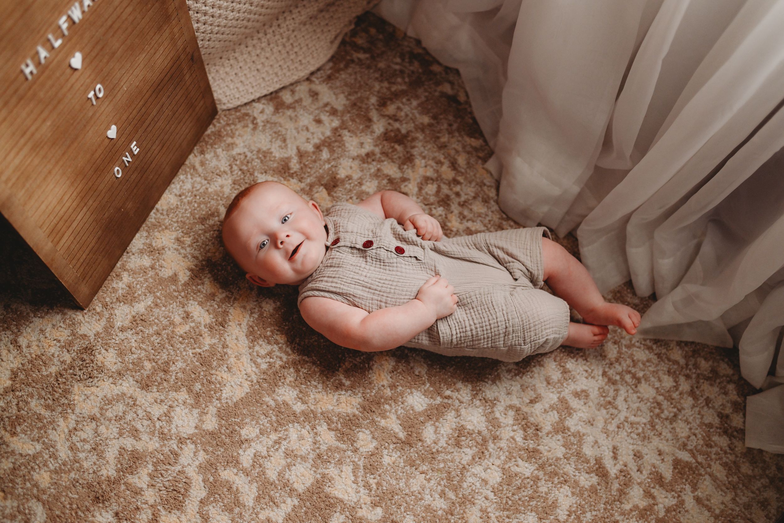 Baby in grey romper lying on patterned carpet beside white curtains.