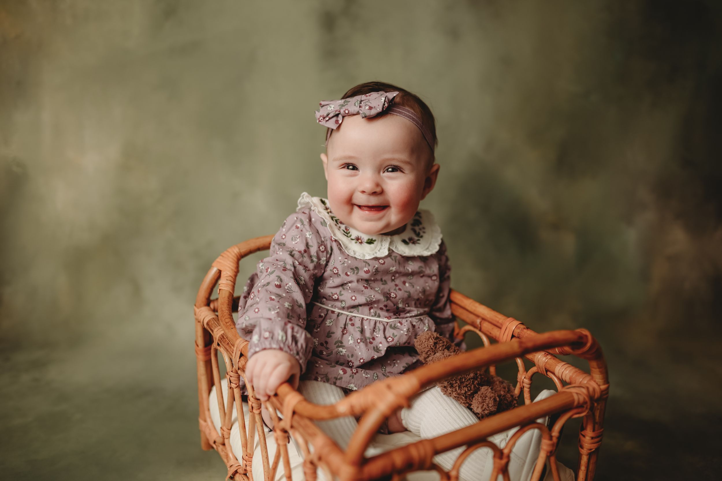 Baby in vintage-style dress sitting in woven rattan chair against muted green backdrop.