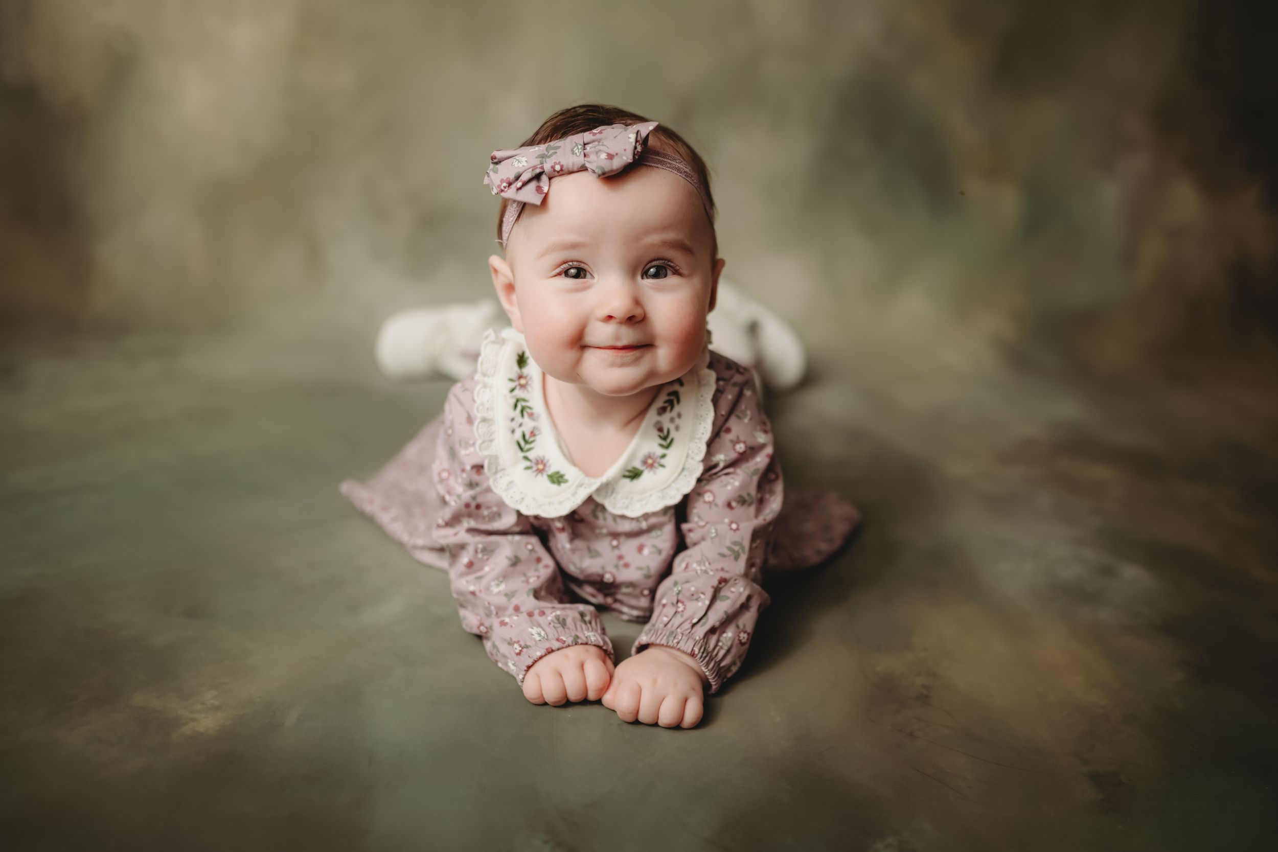 Baby wearing dusty rose dress with lace collar posed against moody green background.