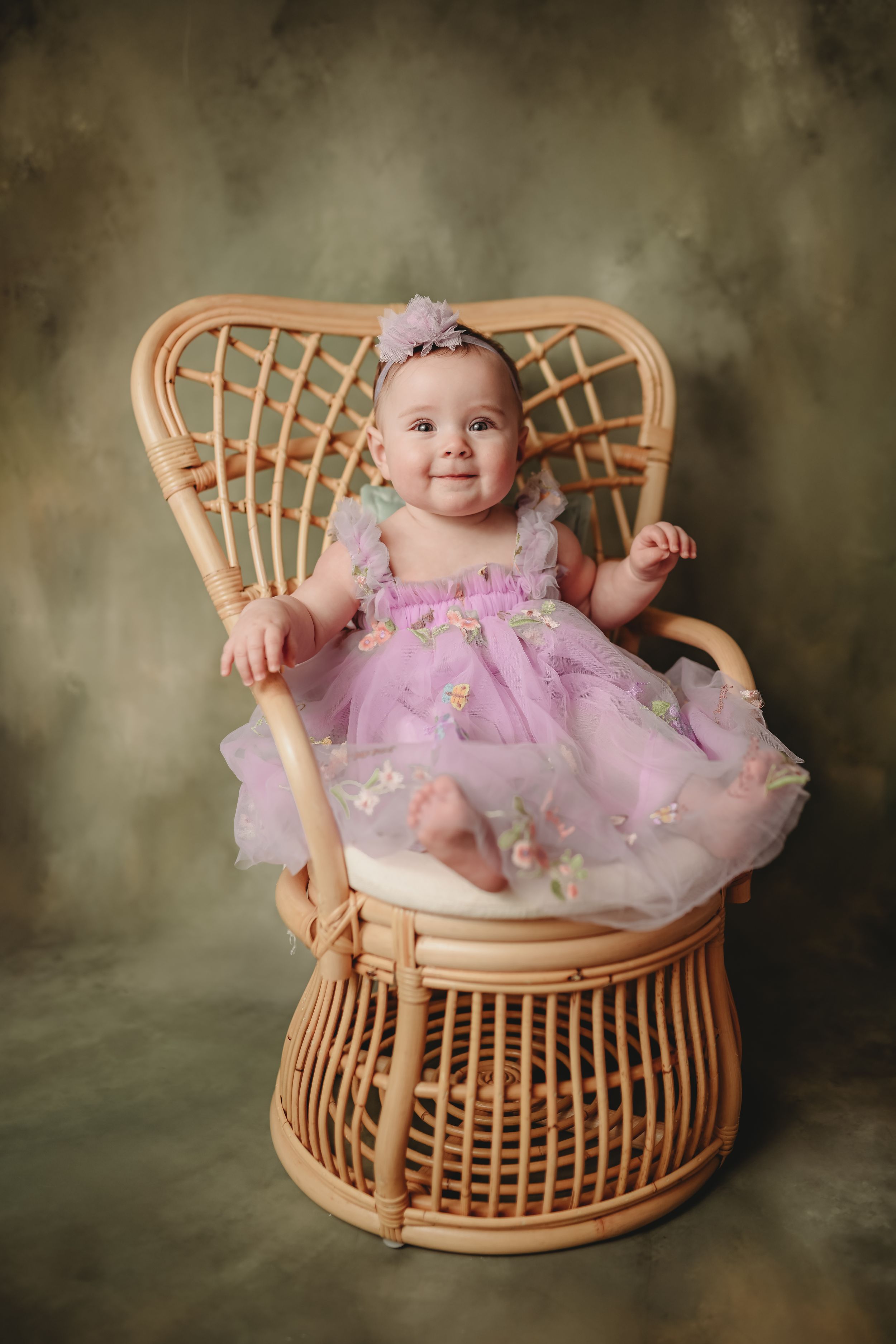 Baby in purple tulle dress sitting in peacock rattan chair against textured backdrop.