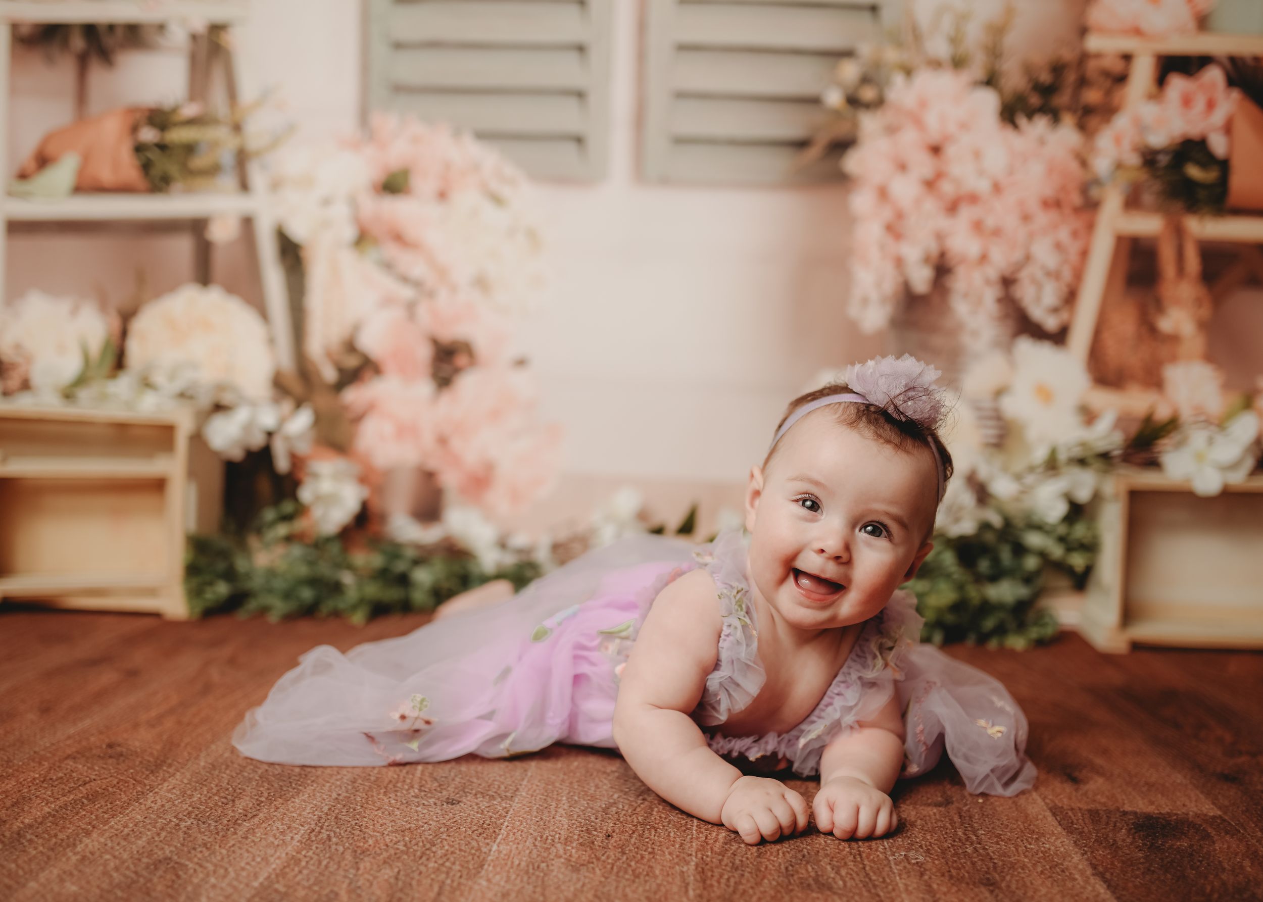 Baby in lavender tulle dress crawling on wooden floor with pink floral backdrop.