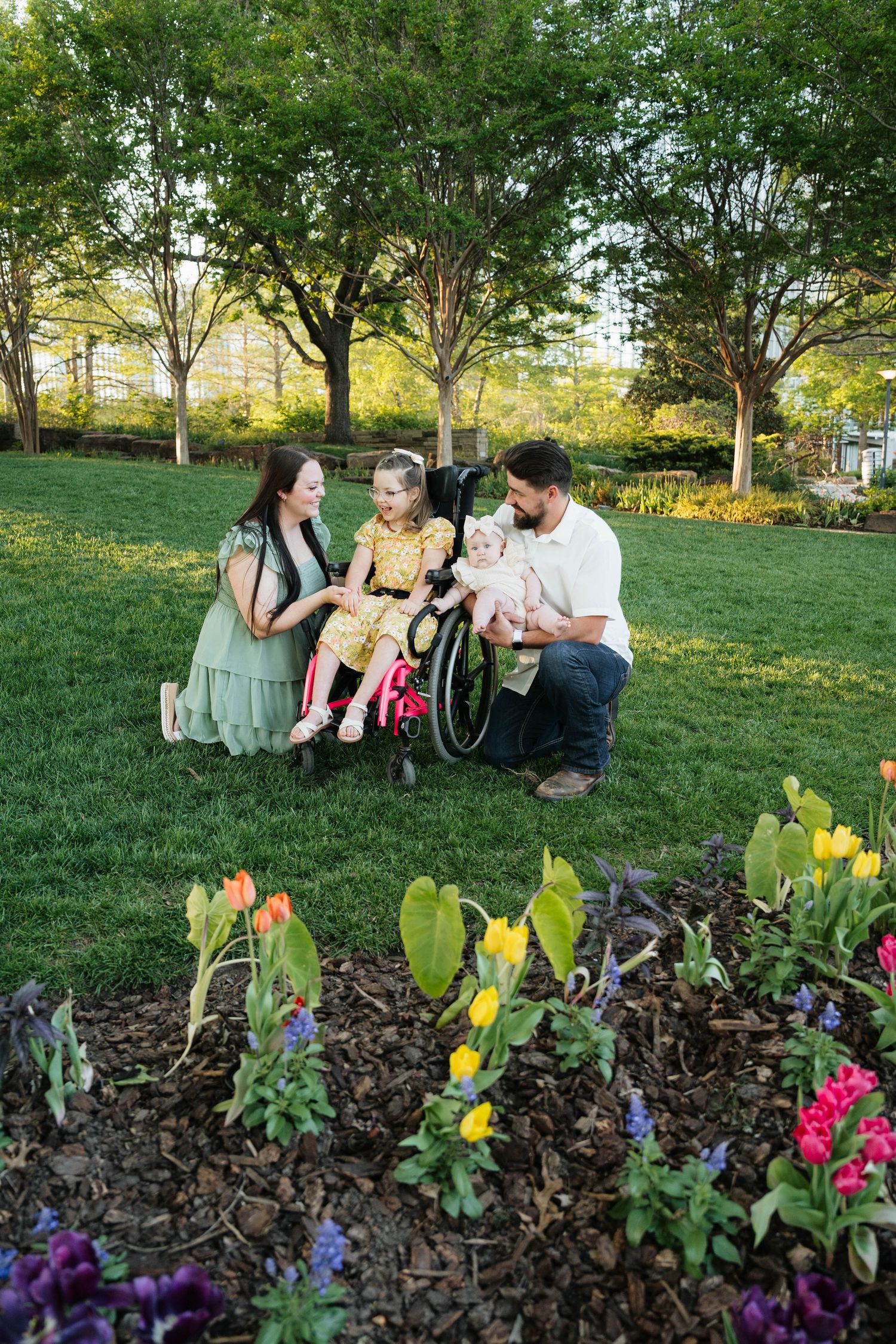Family with two children, one in a pink wheelchair, enjoying a sunny park with colorful tulips in bloom.