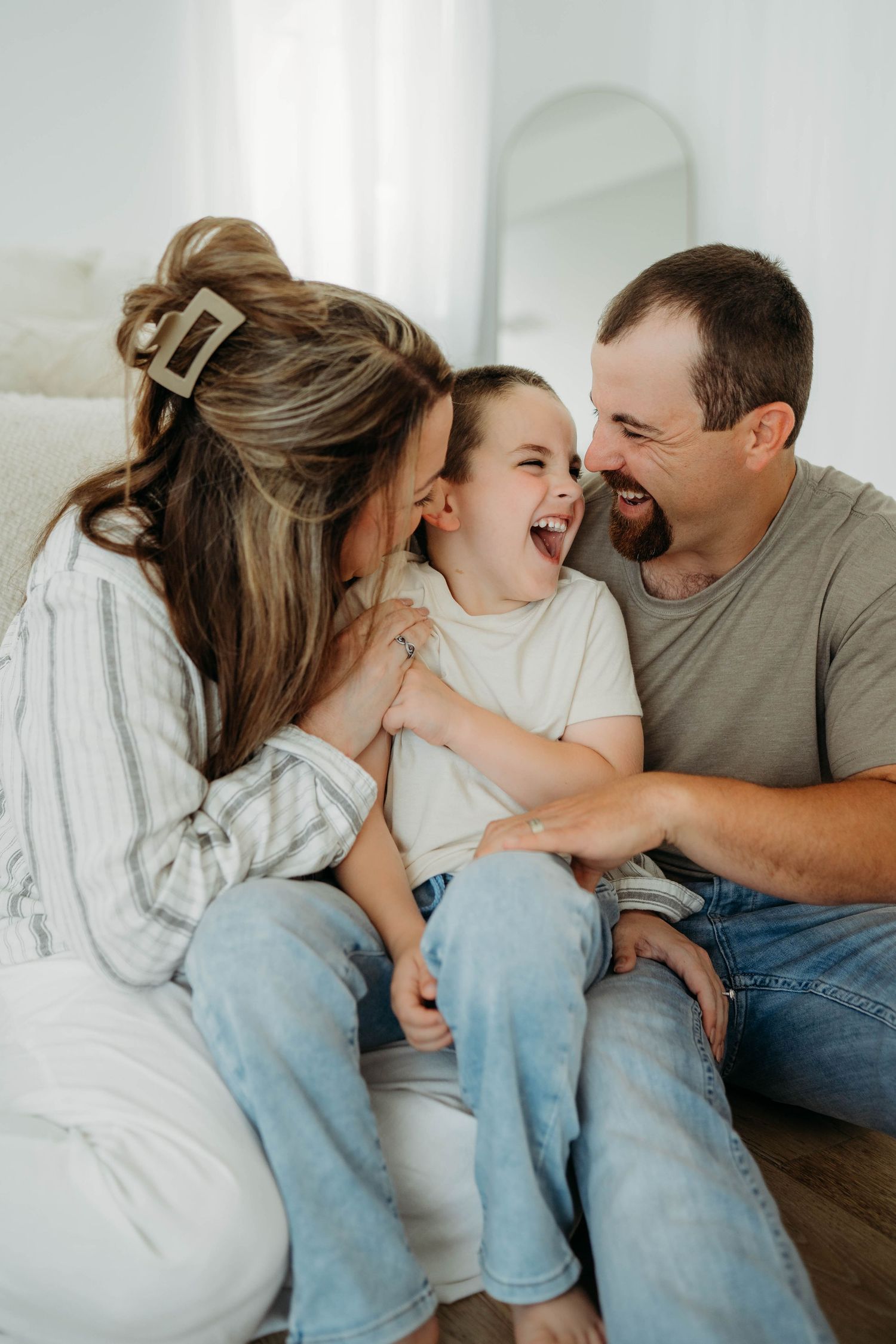A joyful family of three laughing together, with parents leaning in toward their giggling child on a bright white background.