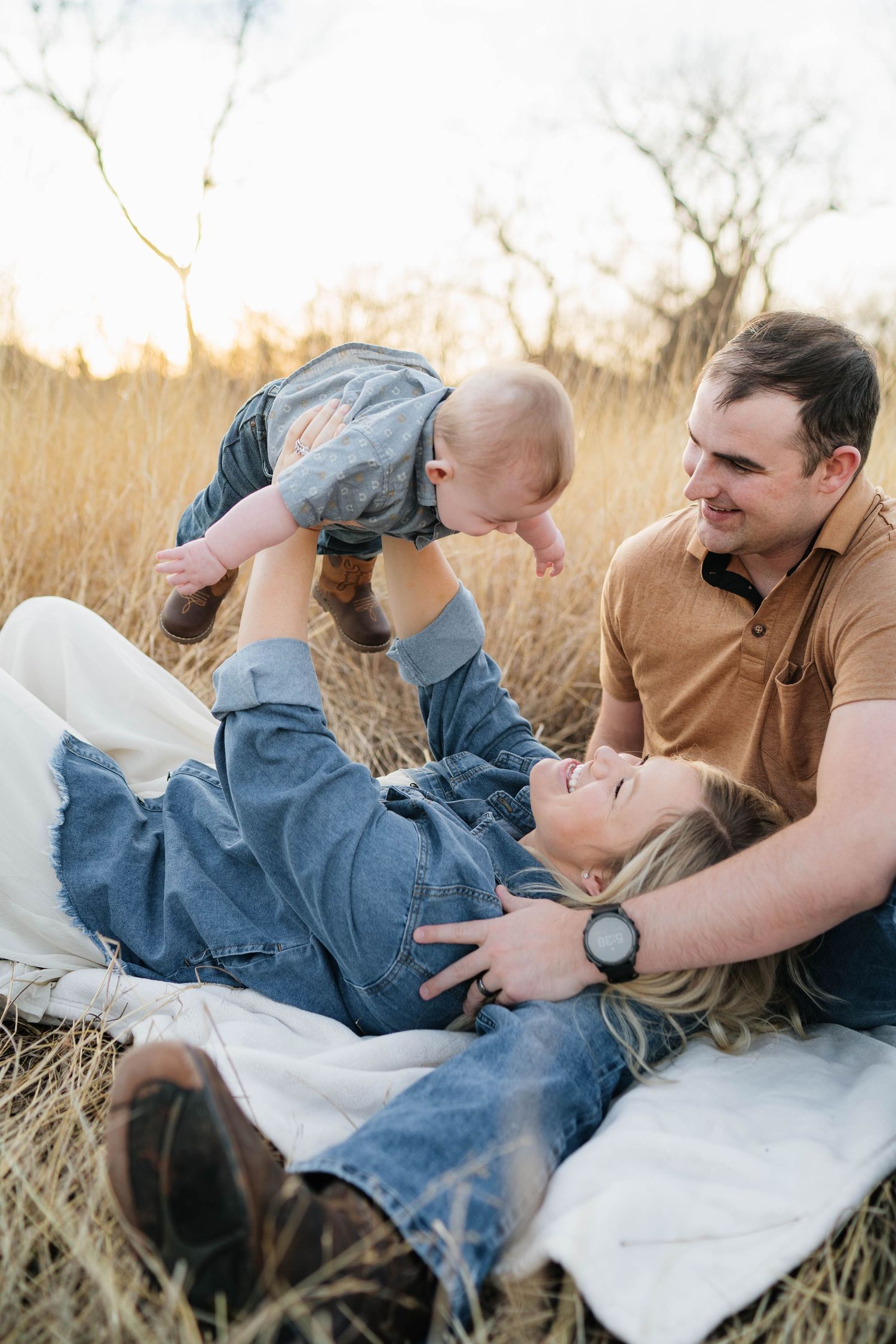 A happy family sits in a golden field at sunset, with a mother lifting her baby while the father watches lovingly.