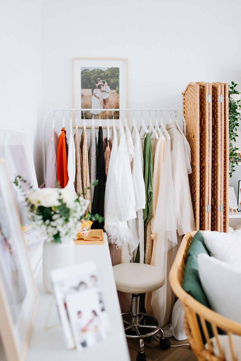 A clothing rack with white and colorful garments next to a wooden room divider and floral arrangement on a desk.