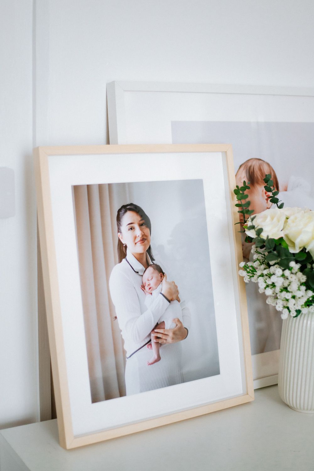 A framed photo sits on a white surface next to a vase of white flowers in a bright, minimalist setting.